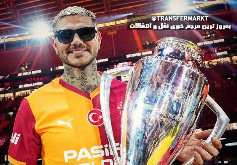 A tattooed man wearing sunglasses and an orange and red striped jersey with a crescent and star emblem poses with a large silver trophy inside a stadium.