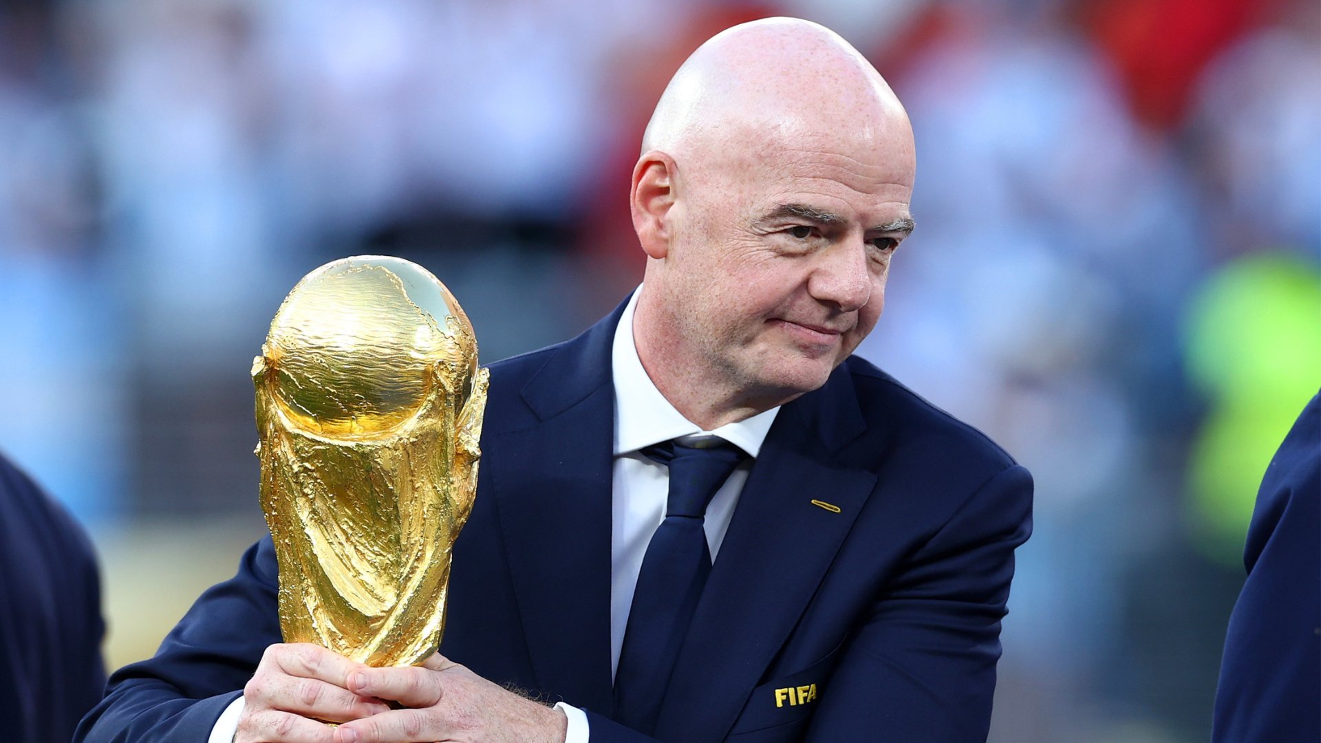 A bald man in a navy suit with a "FIFA" logo pin holds a golden World Cup trophy in front of a blurred crowd.