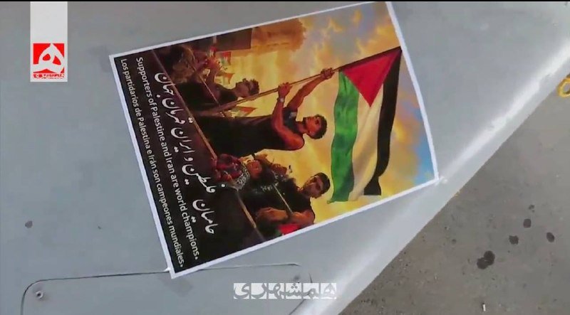 A printed poster on pavement depicts people raising a Palestinian flag, with Arabic text and Spanish text visible in the margins.