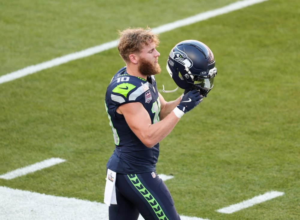 Seattle Seahawks player wearing jersey number 10 holds his helmet on a marked football field.