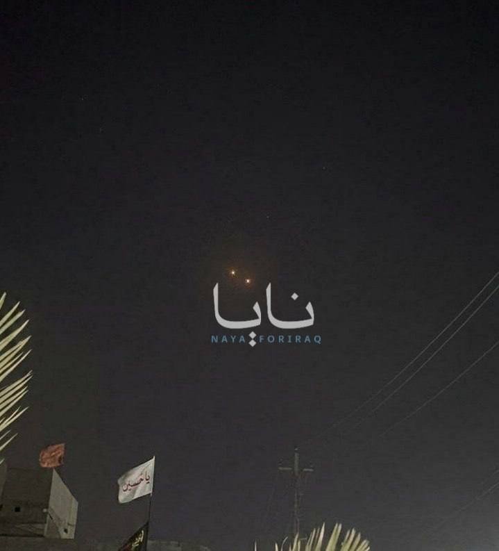 A dark night sky features an overlaid "Naya For Iraq" graphic in Arabic and English, with illuminated rooftop signs and power lines visible below.
