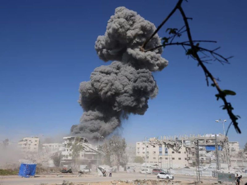 A large plume of dark gray smoke rises above a row of damaged buildings in an urban area under a clear blue sky, with a tree branch visible in the foreground.