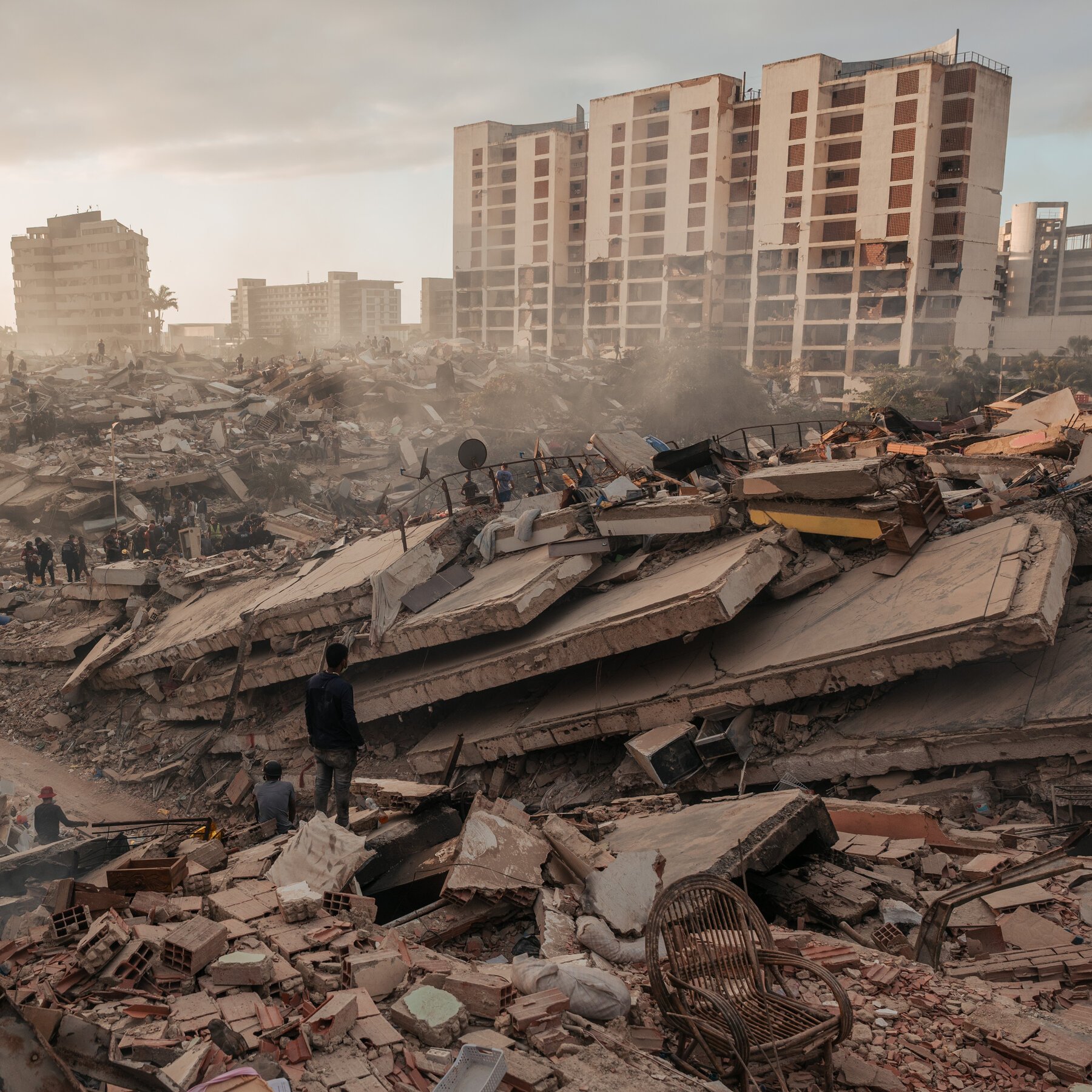Rescue workers sift through massive concrete and brick rubble amid dust, with damaged high-rise apartment buildings visible in the background.