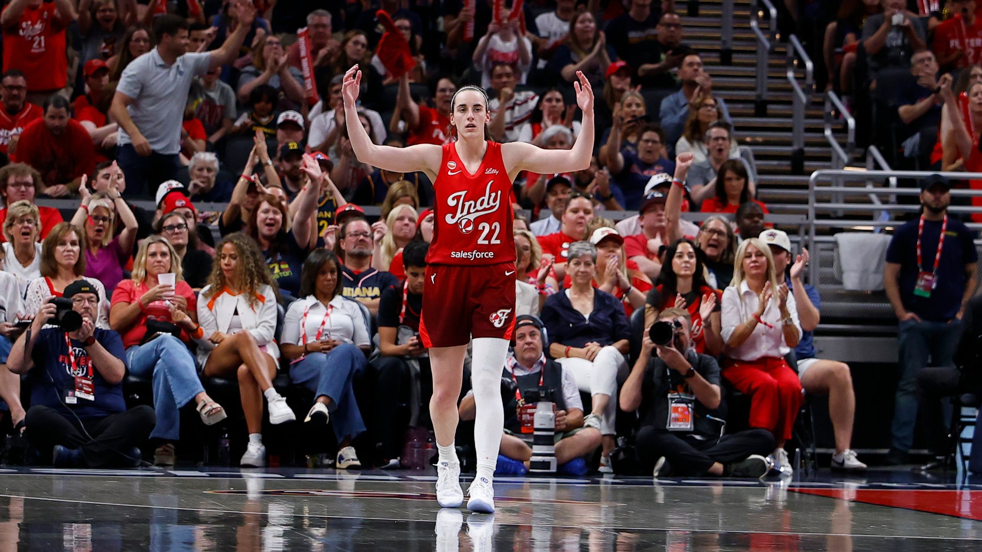 Indiana Fever player wearing jersey #22 raises both arms in celebration as a cheering crowd and seated spectators, including photographers, react behind her at a WNBA game.