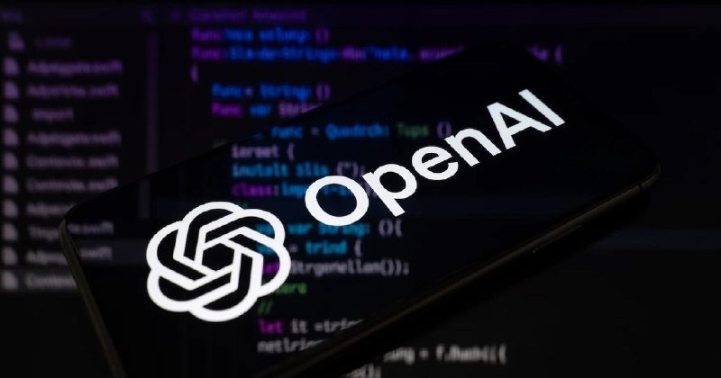A smartphone displaying the OpenAI logo and name rests on a monitor showing lines of programming code.
