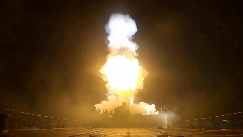 A warship at night fires a large missile upward, producing a bright flame and thick smoke plume against the dark sky.