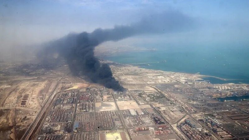 Aerial view of a coastal city with a large plume of dark smoke rising from an industrial area near the shoreline.