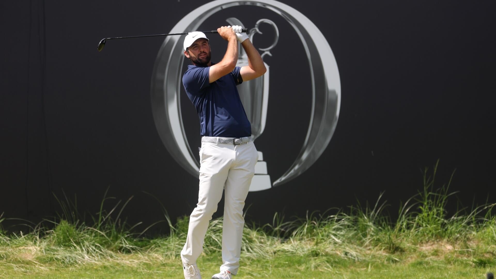 A golfer in a navy shirt, white pants, and white cap completes his swing in front of a dark backdrop featuring a large trophy logo.