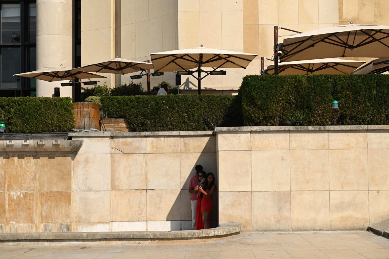 A couple poses for a photo in a shaded alcove of a sandstone building, beneath large patio umbrellas and trimmed hedges on an upper terrace.
