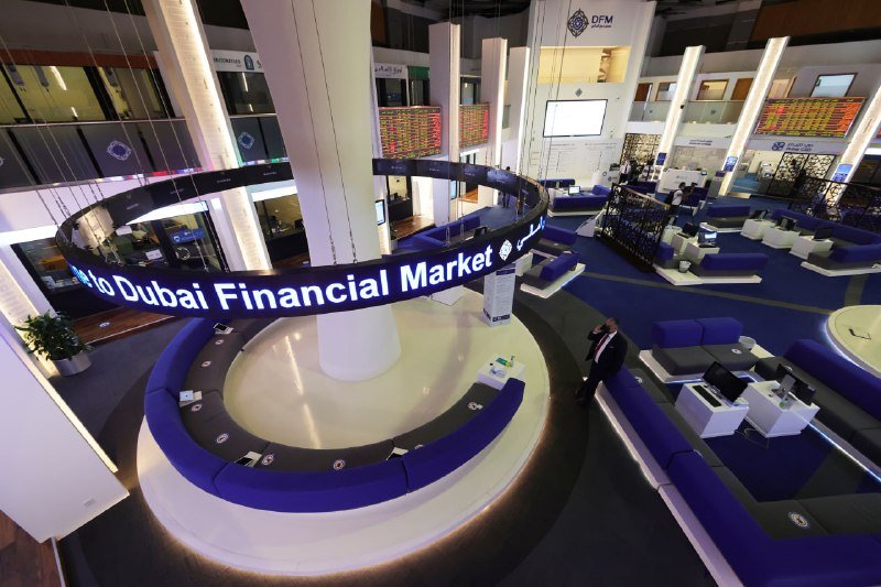 Empty trading floor of the Dubai Financial Market with blue curved seating, digital displays, and a suspended illuminated sign in the center.