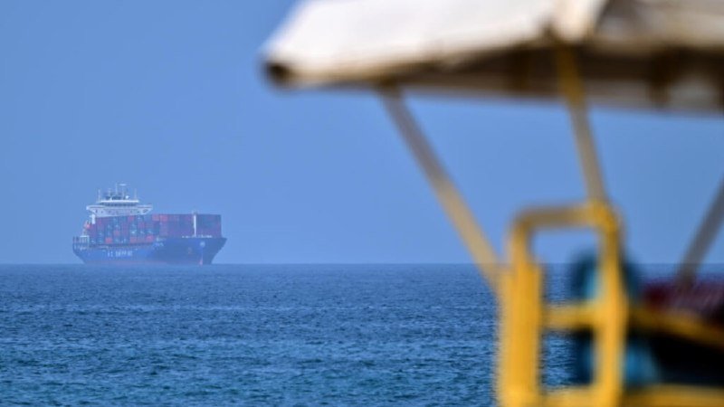 A large blue container ship is visible in the distance on the ocean, framed by the blurred foreground structure of what appears to be another vessel's deck or rig.