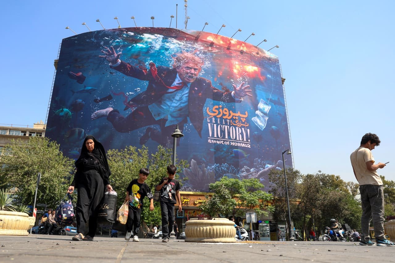 A large outdoor billboard depicts a man in a suit drowning underwater alongside the text "GREAT VICTORY! Strait of HORMUZ," as pedestrians walk past on a sunny street.