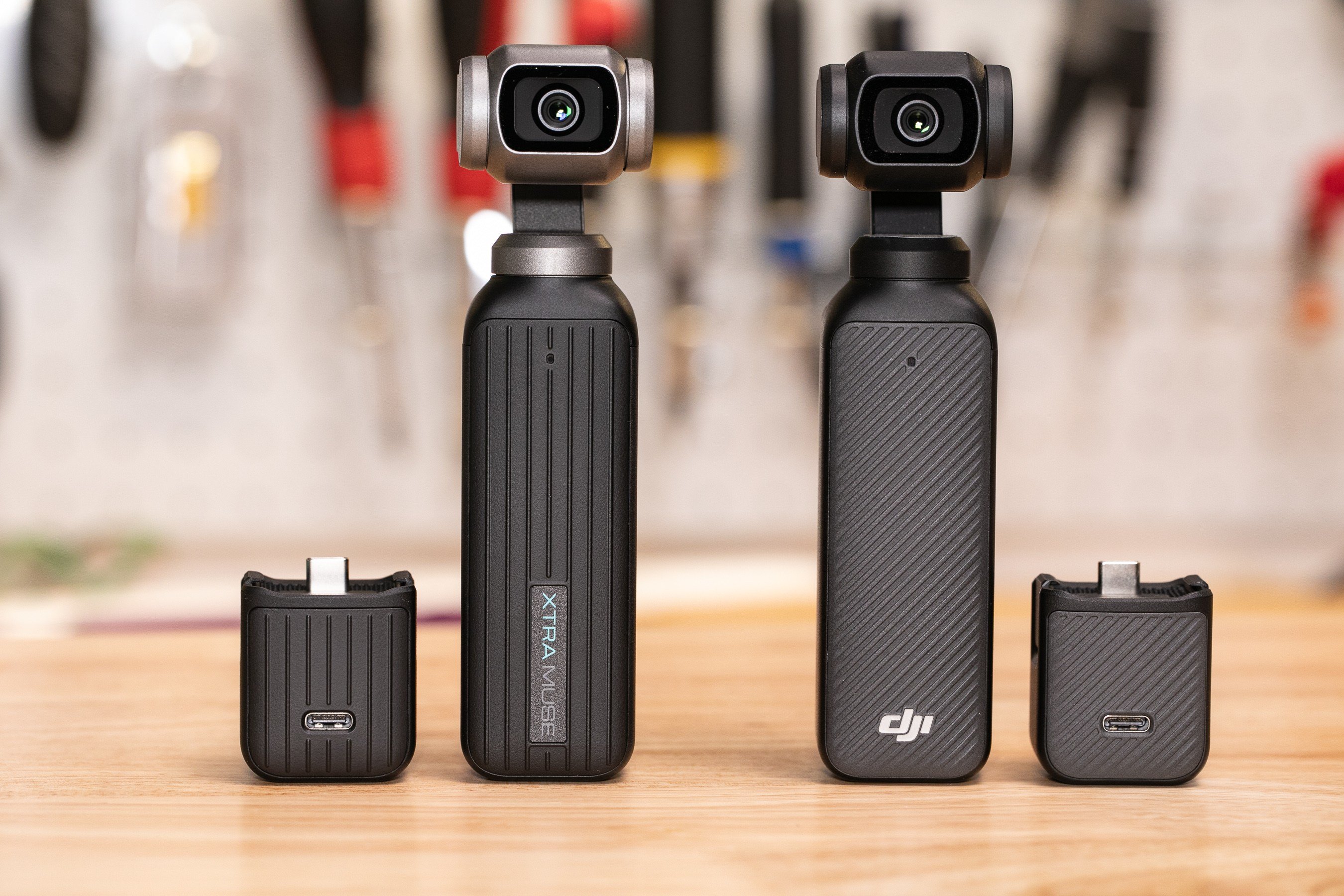 Two compact handheld gimbal cameras and two small adapter units sit side-by-side on a wooden surface, with blurred tools hanging on a wall behind them.