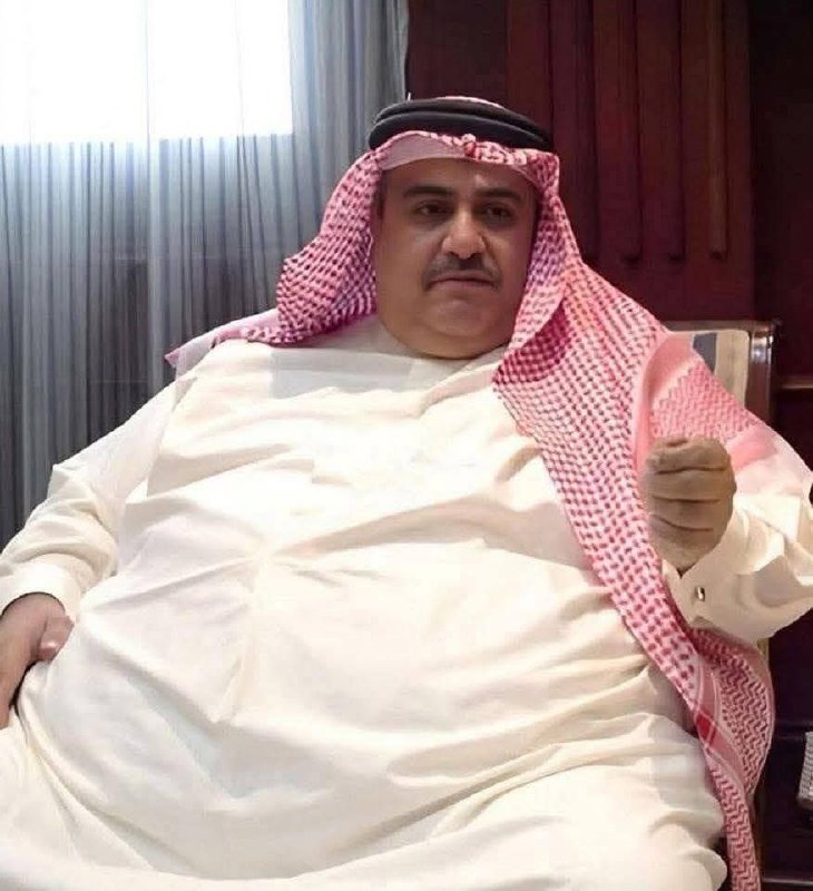 A man wearing a white thobe and red-and-white checkered keffiyeh sits in a chair indoors, gesturing with one hand near wood-paneled walls and curtains.