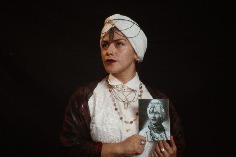 A performer wearing a white turban with jeweled headpiece, ornate necklaces, and a patterned maroon jacket holds up a black-and-white portrait photograph against a black stage background.