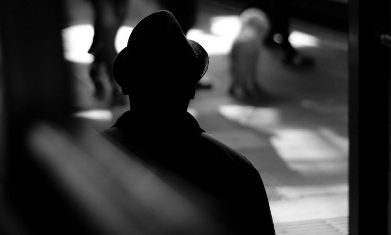A silhouetted figure in a fedora stands in the foreground of a black-and-white street scene, with blurred figures walking in the sunlit background.