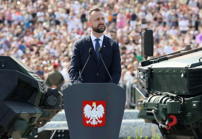 A bearded man in a suit speaks at a podium bearing a white eagle emblem on a red shield, with military equipment and a large crowd visible behind him.