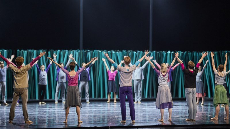 A group of dancers on stage wearing casual clothing stand with their backs to the camera, arms raised in a synchronized pose against a green curtain backdrop.