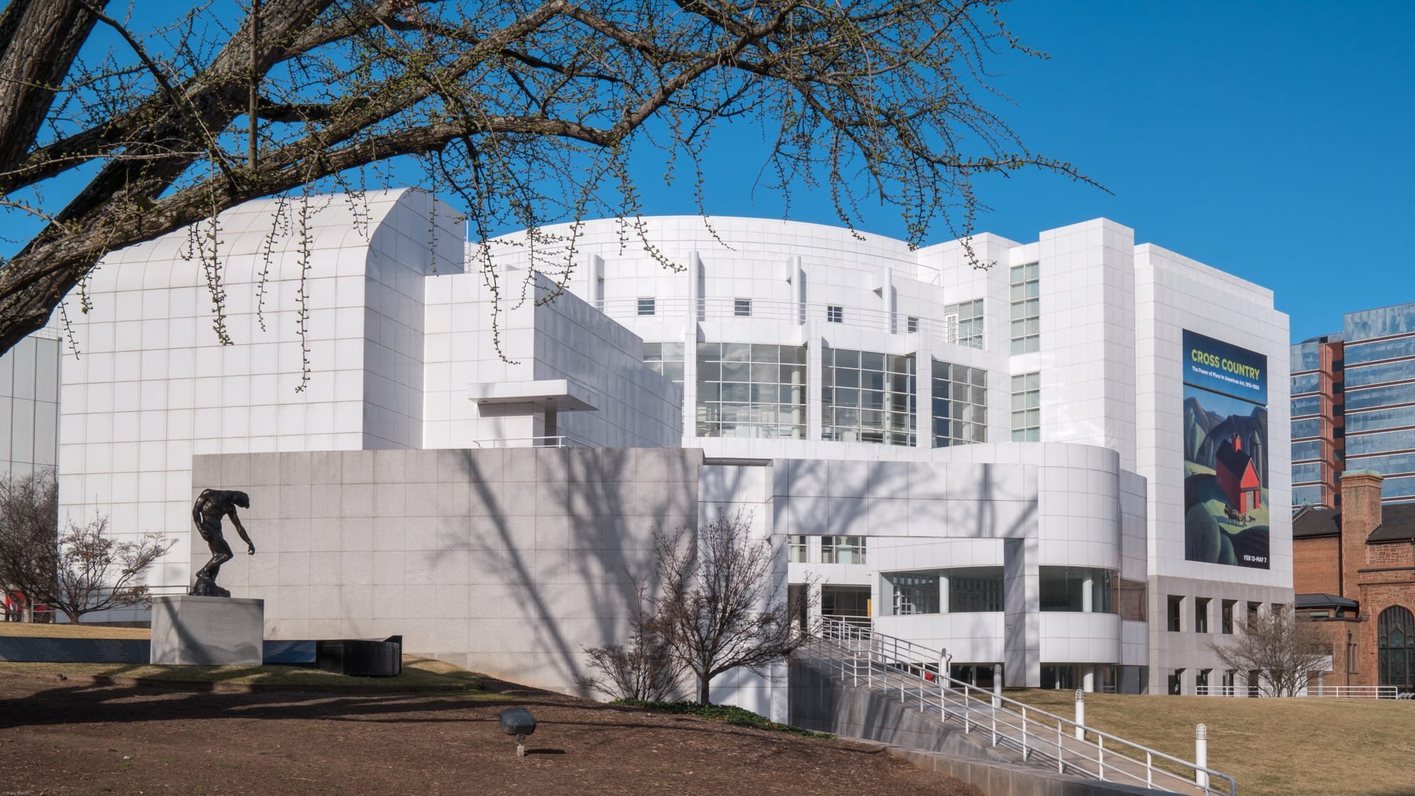 A large white contemporary museum building with curved and angular facades stands under a clear blue sky, featuring a bronze sculpture on a pedestal, bare trees, and a colorful exhibition banner.