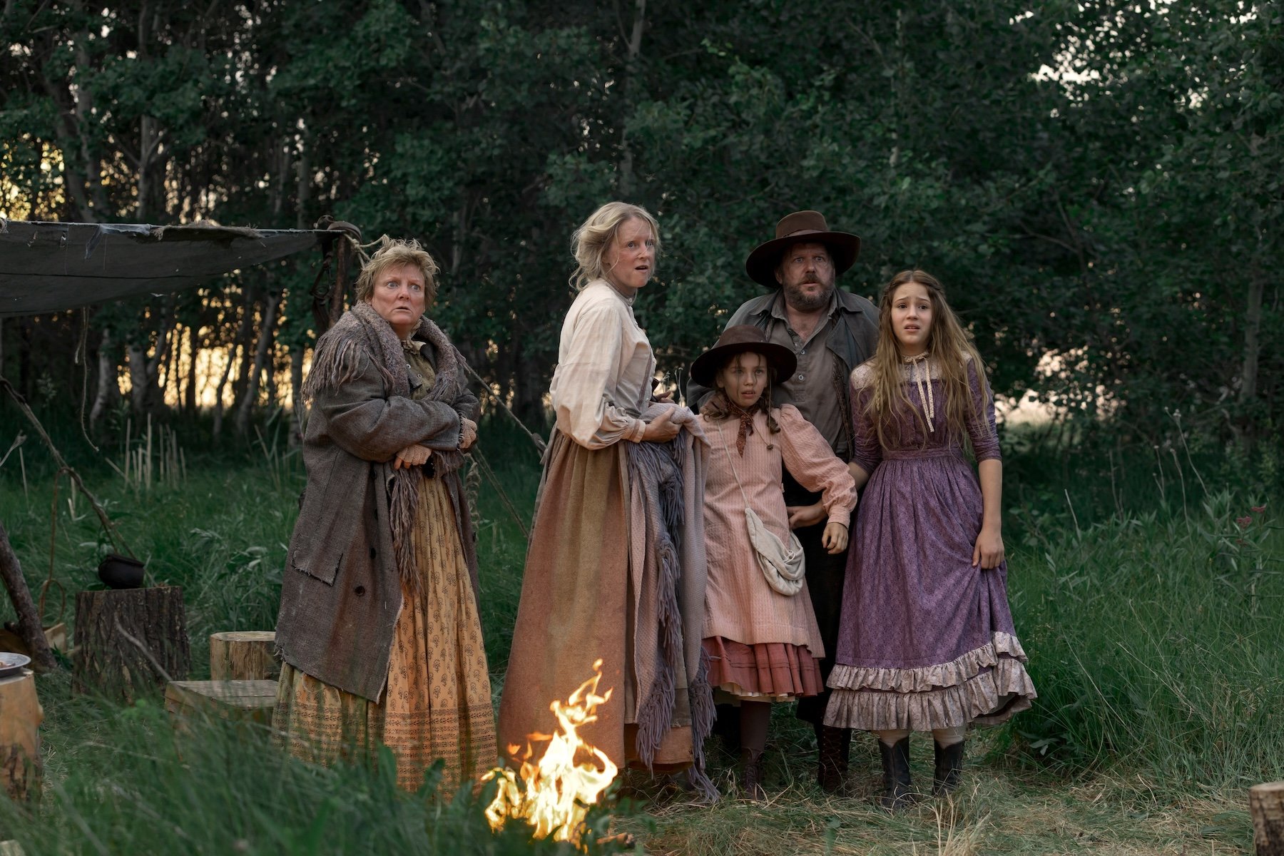 Five people in period frontier clothing stand together near a campfire in a wooded campsite, all looking off-camera with concerned expressions.