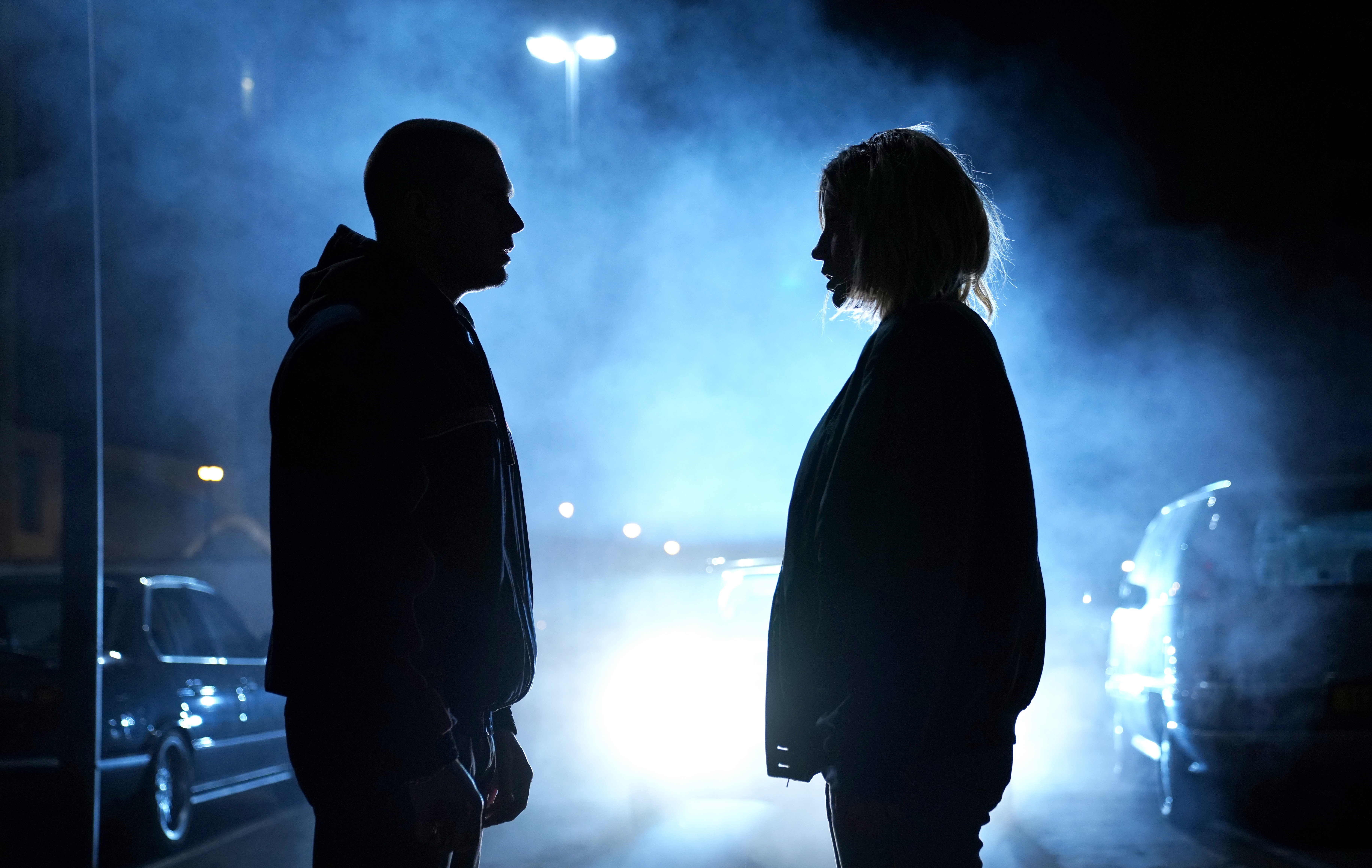 Two silhouetted figures face each other at night on a fog-filled street, illuminated by a streetlamp and the headlights of nearby vehicles.