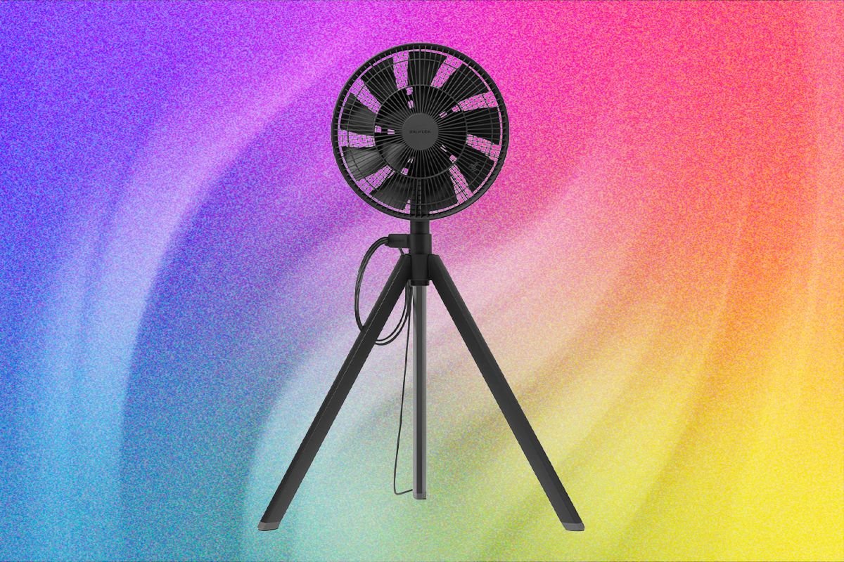 A black tripod-mounted fan stands centered against a vibrant gradient background of purple, pink, orange, and yellow hues.