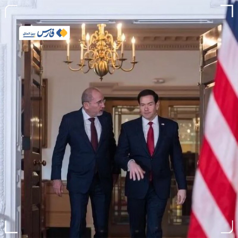 Two men in dark suits and red ties walk through an ornate doorway beneath a chandelier, with an American flag partially visible on the right.