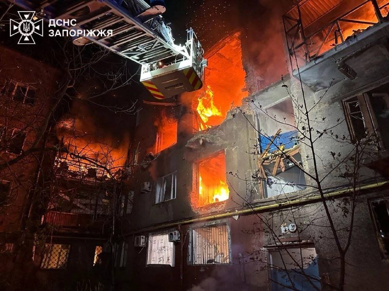 A firefighter in an extended aerial ladder sprays water onto a burning multi-story building at night, with flames visible through broken windows.