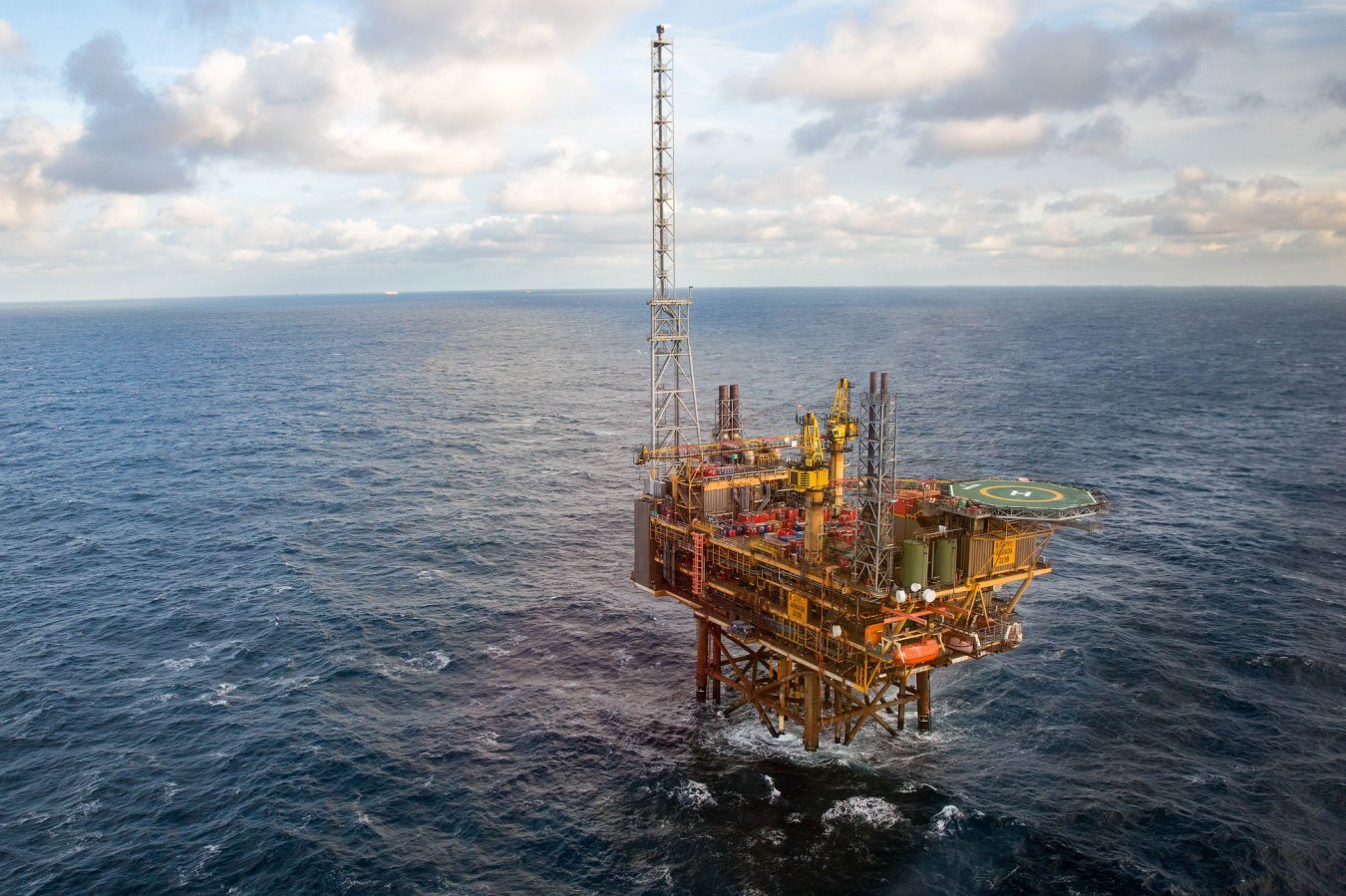 An offshore oil and gas platform with a tall drilling derrick and helicopter landing pad stands in open ocean waters under a partly cloudy sky.