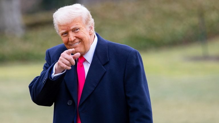 A smiling older man with white hair, wearing a dark navy coat and bright red tie, points toward the camera while standing on a grassy lawn.