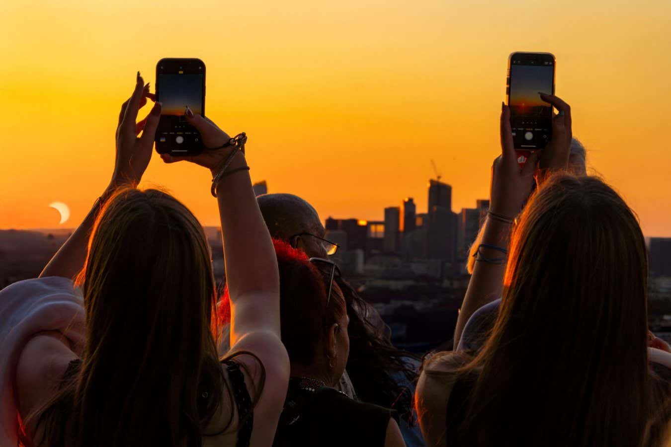 People hold up smartphones to photograph a partial solar eclipse against an orange sunset sky, with a silhouetted city skyline visible in the background.