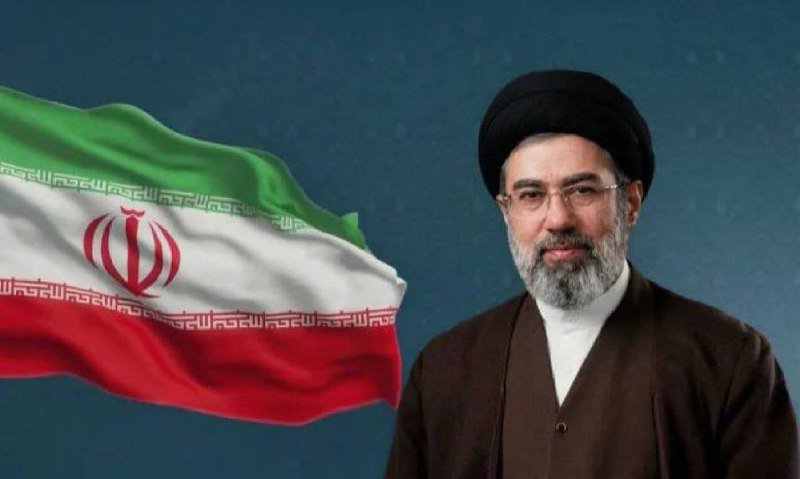 A bearded man wearing glasses, a black turban, and clerical robes stands beside a waving Iranian flag against a dark background.