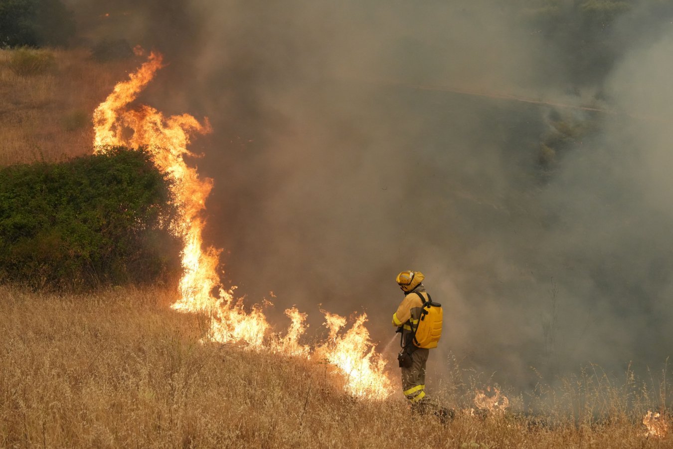A firefighter in yellow gear sprays water on a grass fire burning across a dry, golden hillside, with thick smoke rising into the sky.