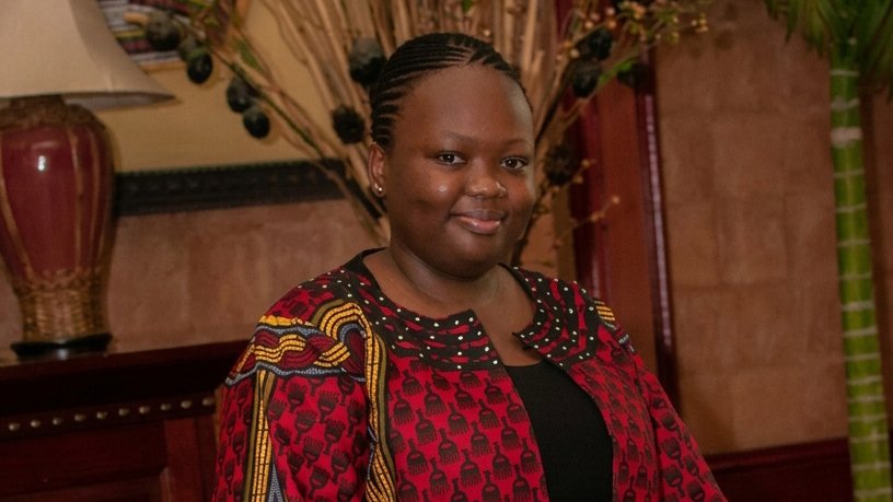 A smiling woman with braided hair wears a red patterned jacket over a black top, posed indoors near a lamp and decorative plants.