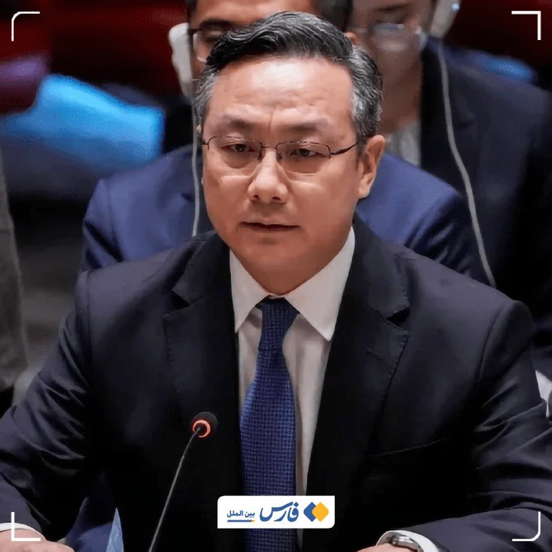 A bespectacled man in a dark suit and blue tie sits at a microphone during a formal meeting, with other attendees visible behind him.