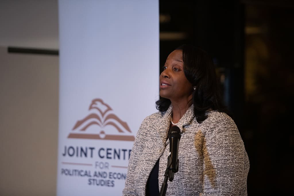 A woman in a tweed jacket speaks at a microphone beside a banner reading "Joint Center for Political and Economic Studies."