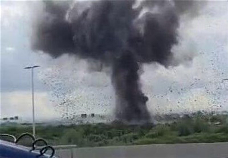 A large plume of thick black smoke rises into the sky, viewed through a rain-spattered window from a roadside with a lamppost and guardrail visible in the foreground.