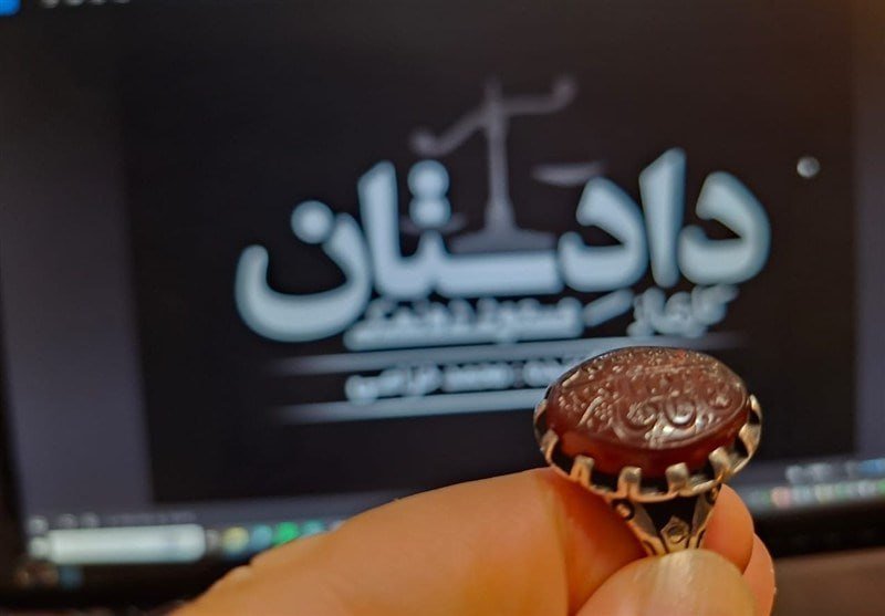 Hand holds a carved reddish gemstone ring in front of a screen displaying Persian text featuring a scale of justice.