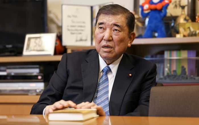 An older man in a dark suit and striped tie sits at a wooden desk with his hands resting on a closed book, speaking in an office setting with shelves behind him.