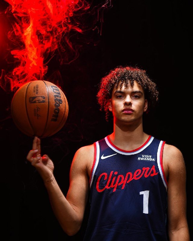 A young basketball player in a navy Clippers jersey labeled "Clippers 1" spins a Wilson basketball on his finger against a dark background with red smoke.