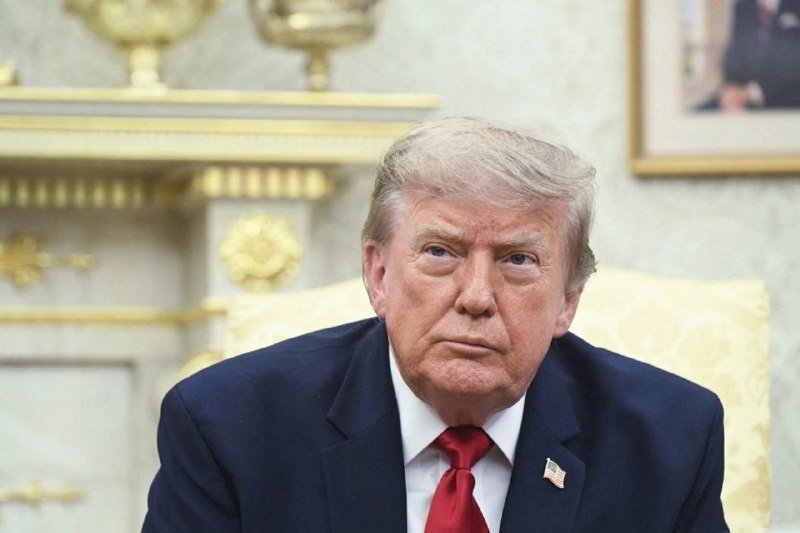 A man with blonde hair wearing a dark suit, white shirt, and red tie sits in an ornately decorated room with gilded decor and framed artwork.