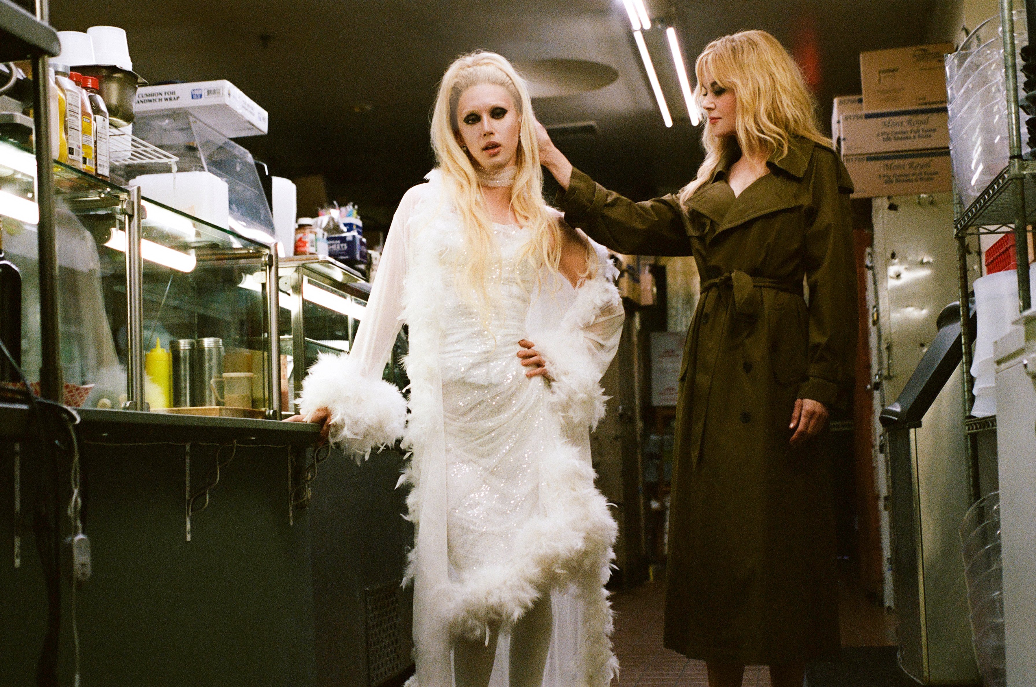 Two blonde women stand in a restaurant kitchen; one wears a feathered white gown while the other, in an olive trench coat, touches her hair.