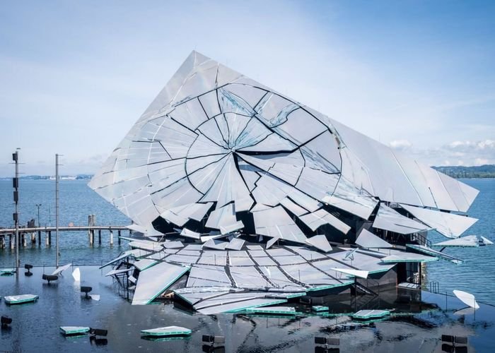 A shattered glass sculpture rests on a flooded platform beside a lake, with broken panels scattered across the water's surface under an overcast sky.