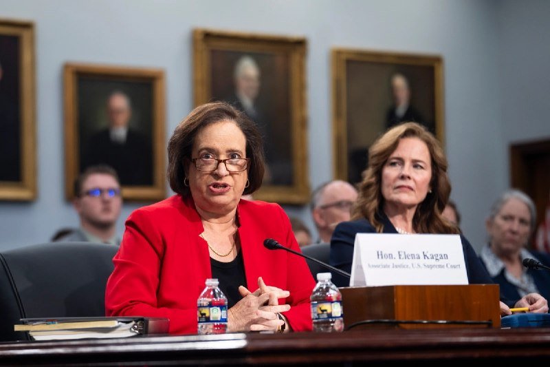 A woman in a red jacket speaks at a hearing table, with a name placard reading "Hon. Elena Kagan, Associate Justice, U.S. Supreme Court" visible in the foreground.