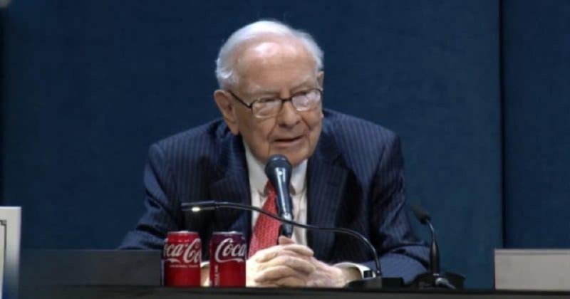 Warren Buffett speaks into a microphone with two Coca-Cola cans on the podium before him.