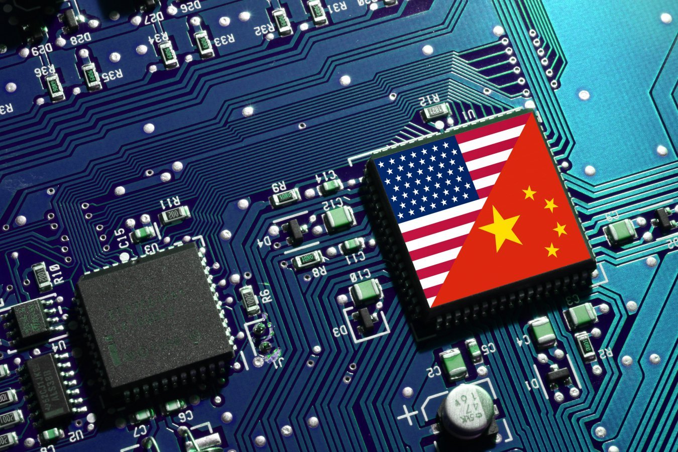 A computer chip bearing the flags of the United States and China rests on a blue printed circuit board populated with microchips, resistors, and capacitors.