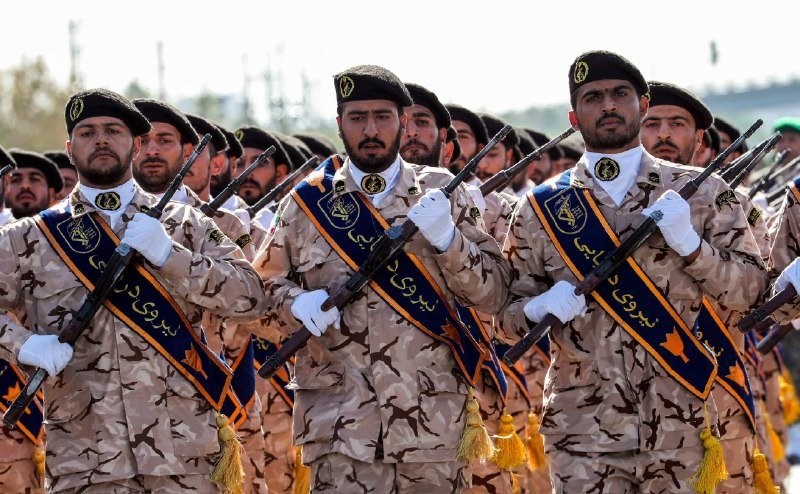 Soldiers in camouflage uniforms and black berets march in formation while carrying rifles and wearing blue and yellow sashes.