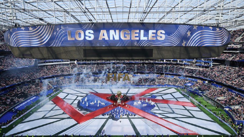 A packed stadium features a "LOS ANGELES" banner above a FIFA ceremony display, with performers arranged around a golden trophy on a stage decorated in red, white, and blue patterns.