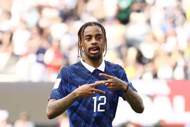 A soccer player wearing a blue jersey with the number 12 makes hand gestures in front of a blurred crowd of spectators.