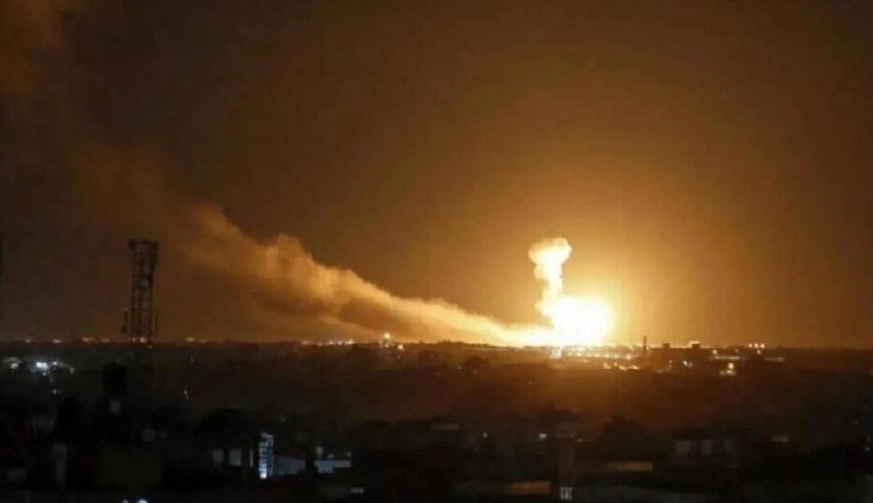 A large explosion erupts with a bright orange fireball and trailing smoke plume over a darkened cityscape at night.